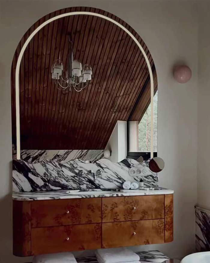 Połokrągłe podświettlane lustro LED w łazience inspirowanej stylem mid-century modern z drewnianą zabudową czeczota i marmurem