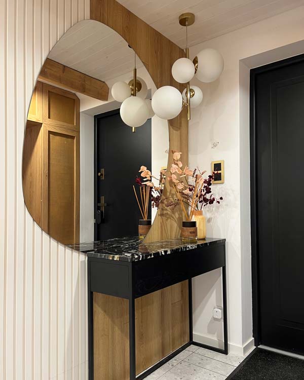 Nieregularne lustro Rocoo zawieszone asymetrycznie nad konsola w nowoczesnym wiatrołapie z drewnianą szafą i przeszklonymi  drzwiami